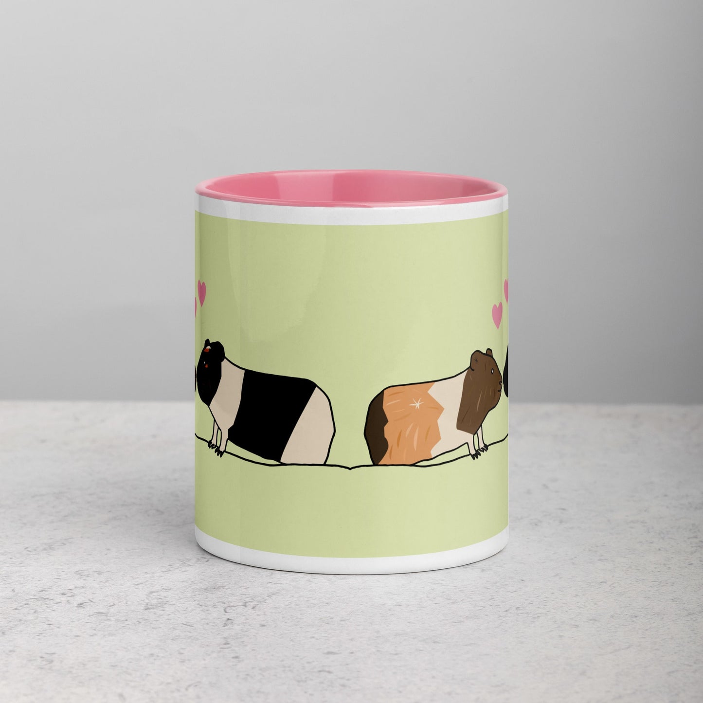 Front view of green ceramic mug with two cute guinea pigs, one black and white, the other brown and white, against a colorful pink interior.