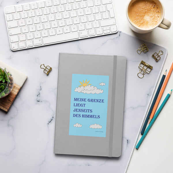 Front cover of the hardcover notebook with "Meine Grenze liegt jenseits des Himmels" on a desk beside a computer and mug, creating a cozy lifestyle setting.