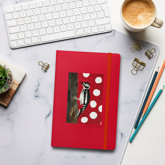 A red hardcover notebook with an elastic closure and a printed woodpecker illustration on the cover, arranged on a white marble desk with a keyboard, coffee cup, binder clips, pencils, and a small succulent.