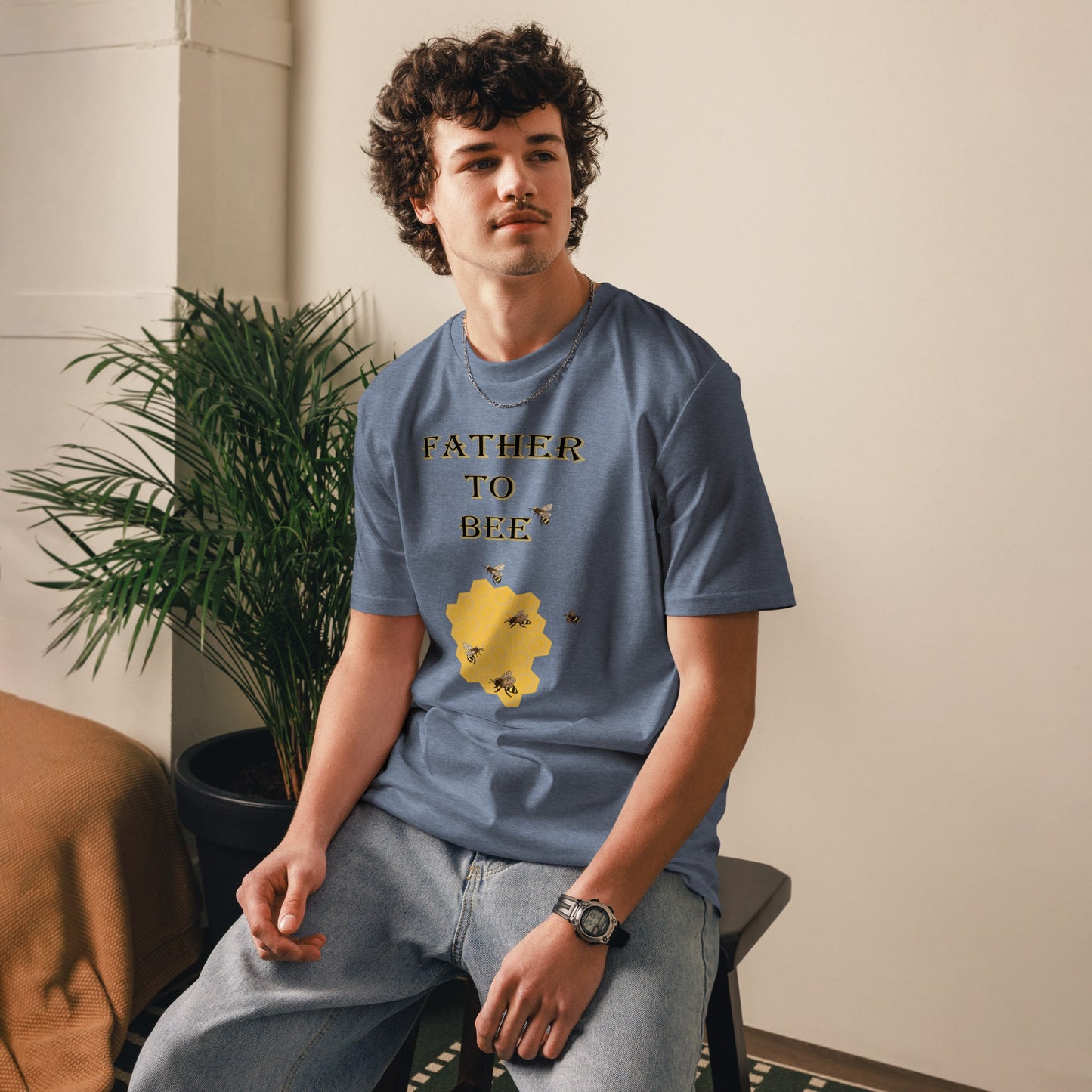 Man wearing a dark heather blue “Father to Bee” organic cotton t-shirt at a slight front angle, standing in a cozy room setting.