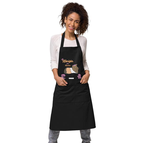 A woman models a black apron with a colorful guinea pig print and “Königin der Küche” in vibrant pink lettering.