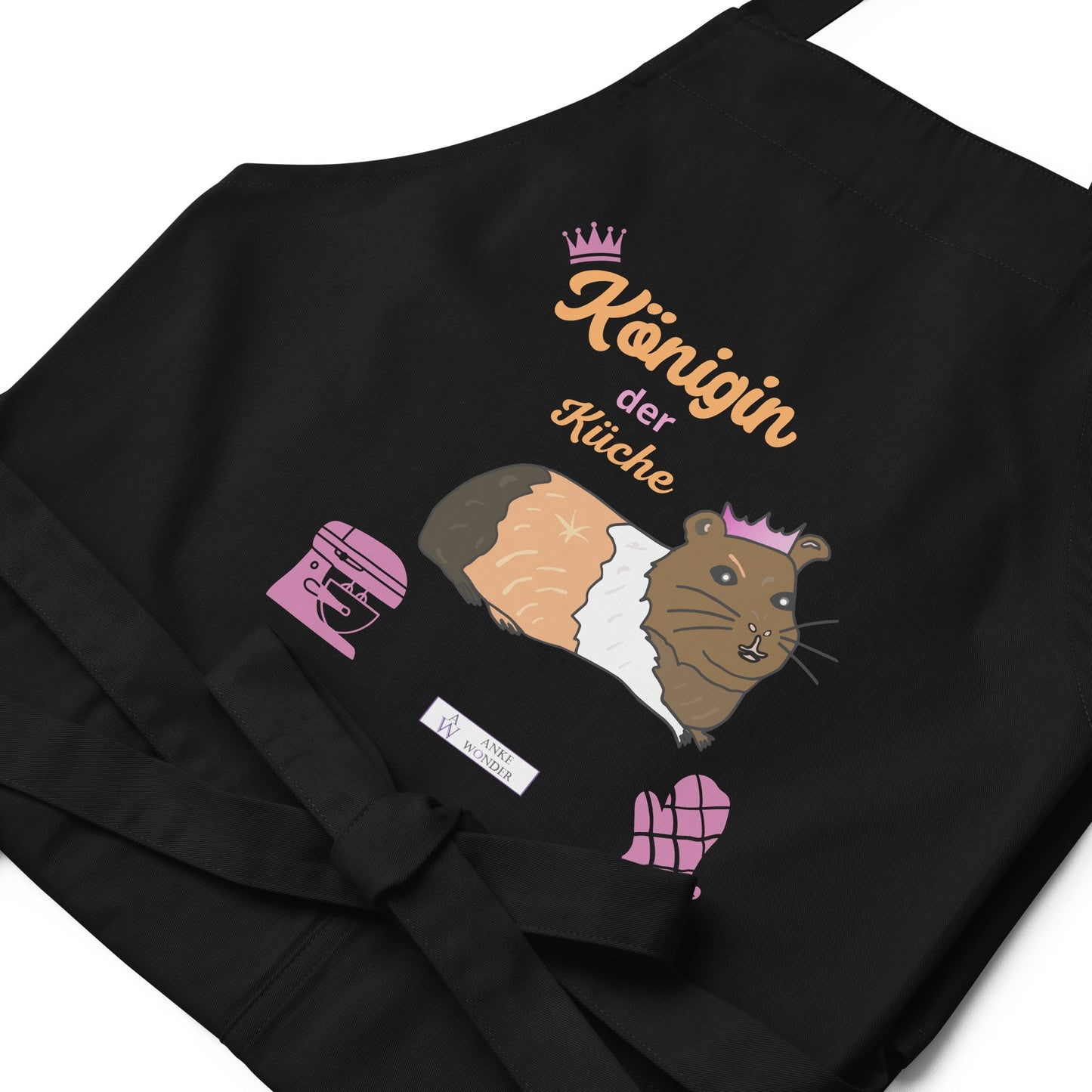 A flat-lay image of the black apron highlighting the detailed guinea pig illustration and the “Königin der Küche” lettering as well as the Anke Wonder branding.