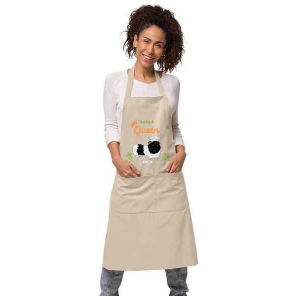 A woman wears a beige apron with a cute guinea pig chef design and the words 'Salad Queen' in orange and green.