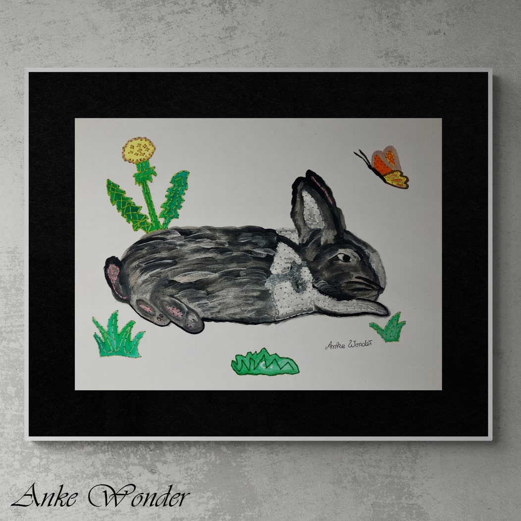 A mixed-media artwork titled "Rabbit in Meadow" by Anke Wonder, featuring a black and white rabbit resting in a green meadow with embroidered dandelions, grass, and a butterfly. The piece combines acrylic painting with hand-embroidery for added texture and depth, framed with a black matting and displayed against a neutral wall.