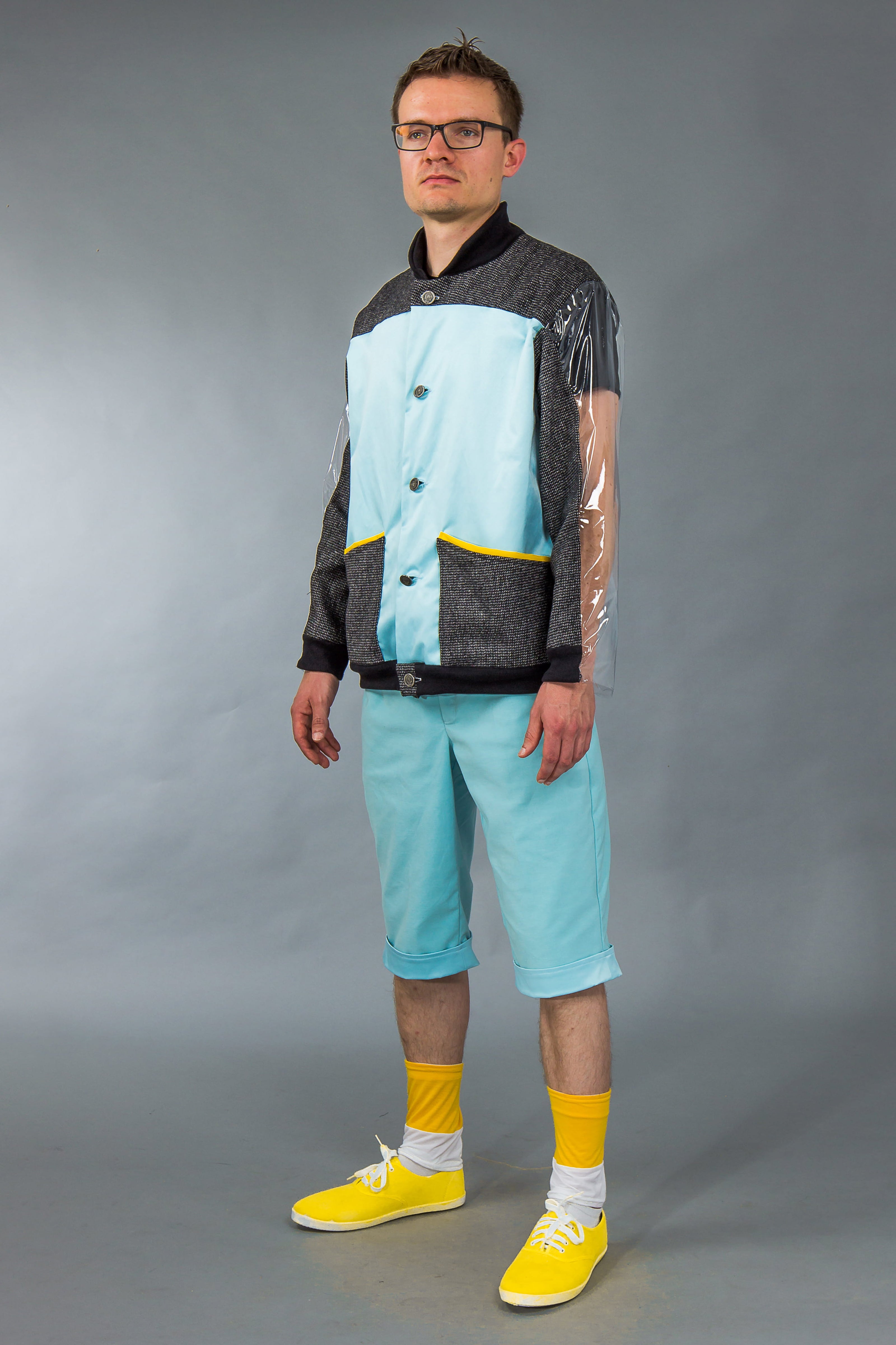 Man wearing a stylish outfit with a gray jacket, light blue shirt, shorts, and yellow shoes on a gray background