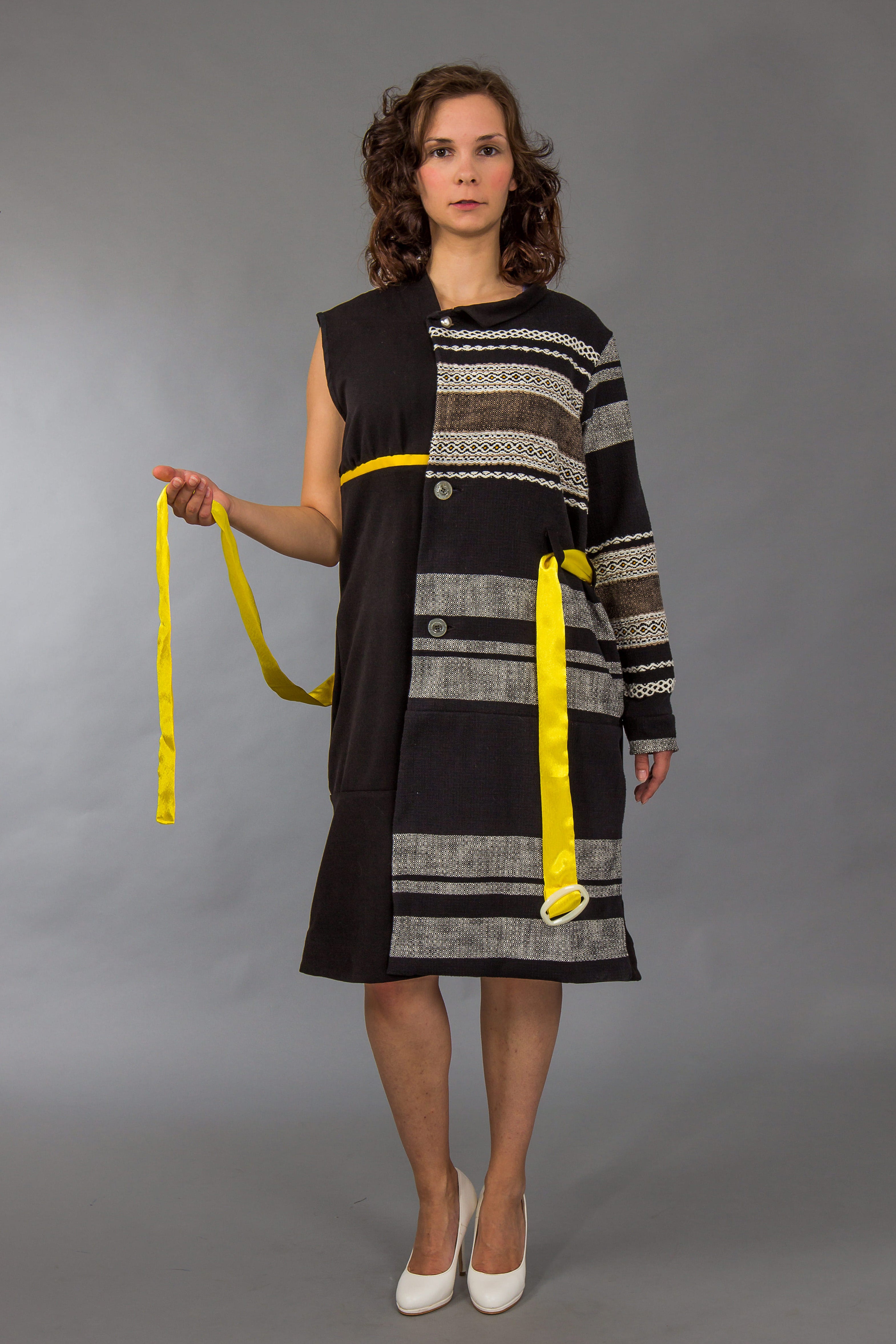 Woman wearing a black and gray patterned dress with yellow accents, holding a measuring tape against a gray background.