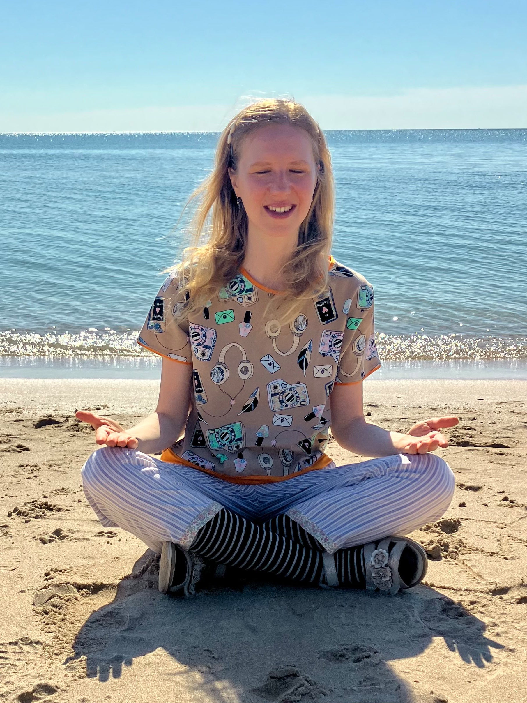 Anke Wonder sitting on the beach with a patterned shirt and striped pants, smiling in a meditation pose.