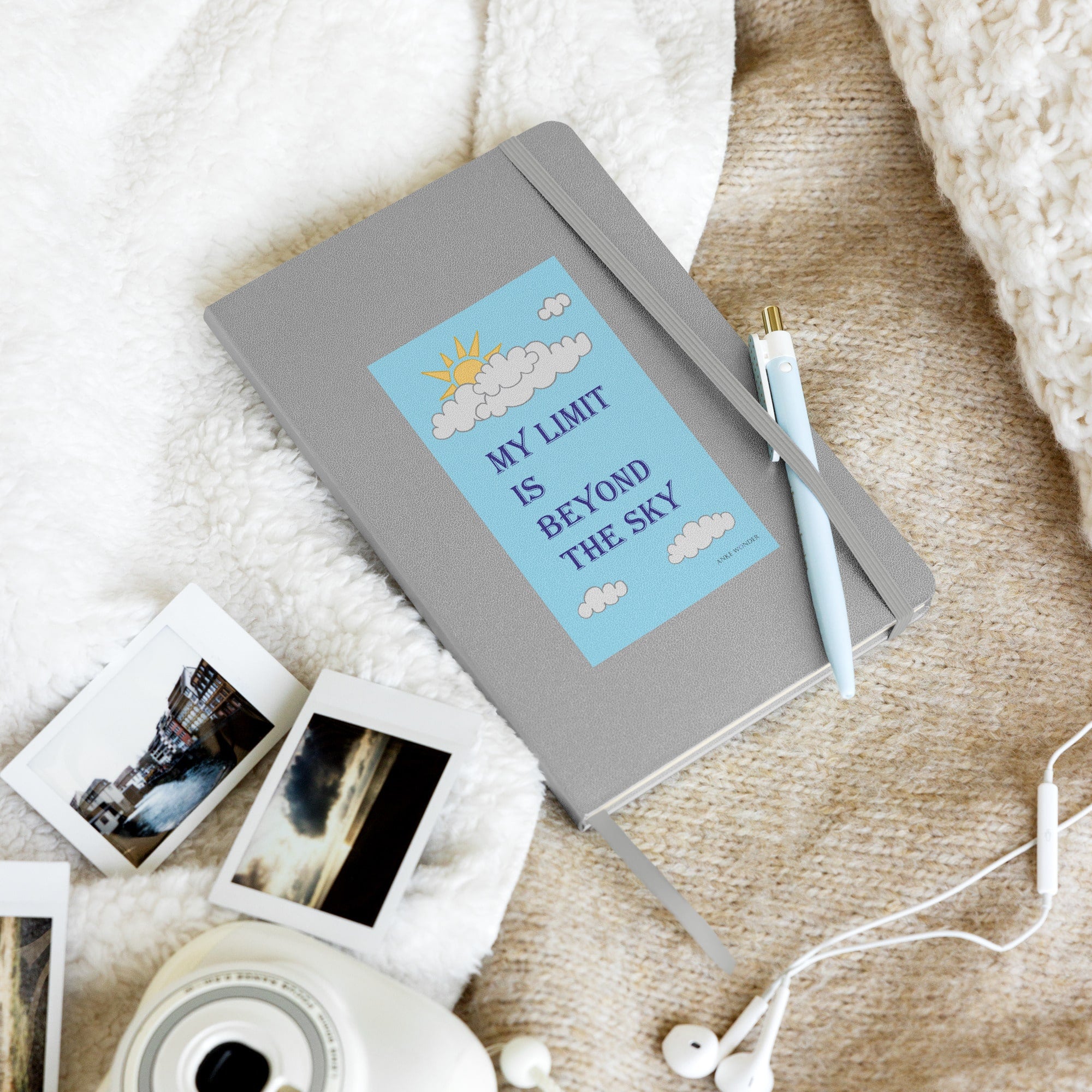 Front cover of the hardcover notebook with "Meine Grenze liegt jenseits des Himmels" on a blanket beside a photos and headphones, creating a cozy lifestyle setting.