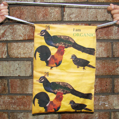 Brown tote bag with peacock, bird and chicken and the words 'I#m Organic' in front of a wall.