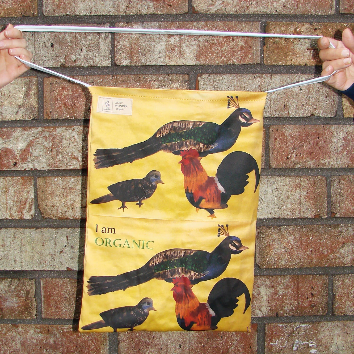 Brown tote bag with peacock, bird and chicken and the words 'I#m Organic' in front of a wall.