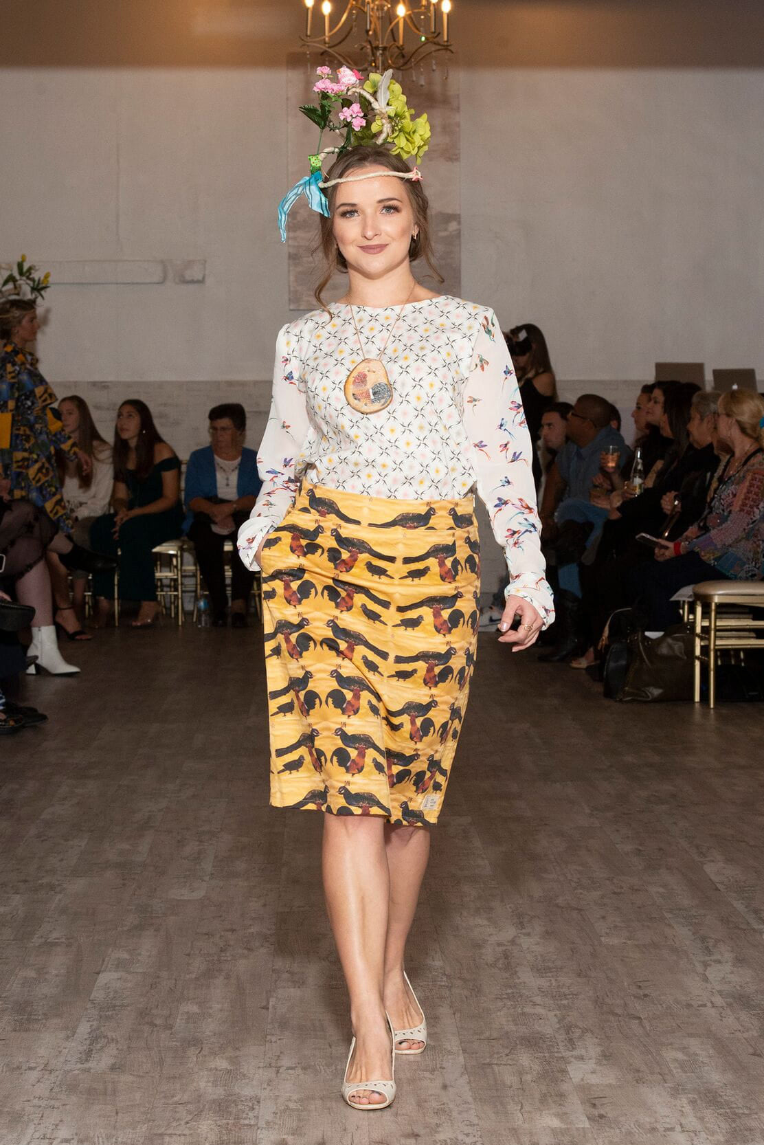Model walking on a catwalk wearing a floral top and patterned skirt with an audience in the background.