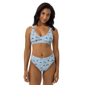 A woman with medium skin tone and dark wavy hair wears a high-waisted, eco-friendly bikini with a light blue background featuring a playful manatee and turtle print. She stands confidently against a white backdrop, showcasing the sustainable swimwear made from recycled materials with UPF 50+ protection.