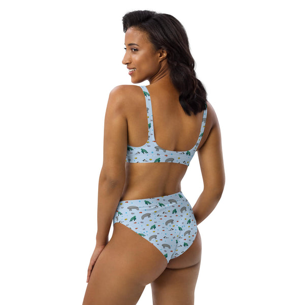 A woman poses in a recycled fabric bikini, turned slightly to the back left, showcasing the curve-hugging fit and ocean-themed print.