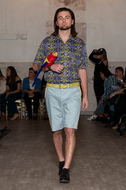 Model walking on a runway wearing a patterned shirt and light shorts, holding a parrot.
