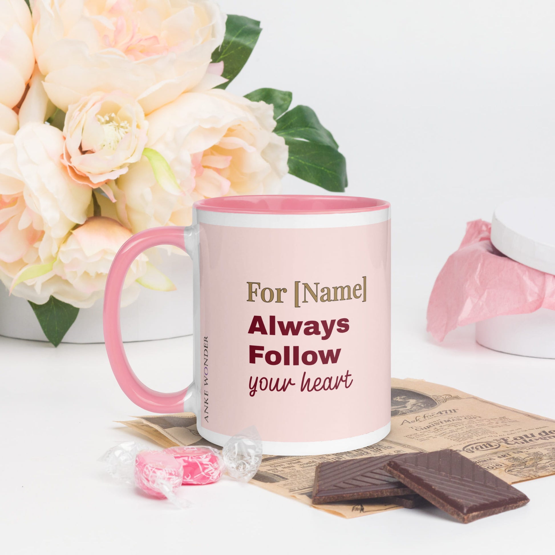 Personalized mug with pink handle and text, surrounded by flowers and chocolates on a light background
