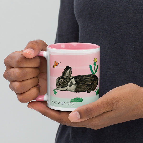 A ceramic mug with a pink rim, handle, and interior featuring a hand-painted bunny, dandelion, and butterfly - designed by Anke Wonder.