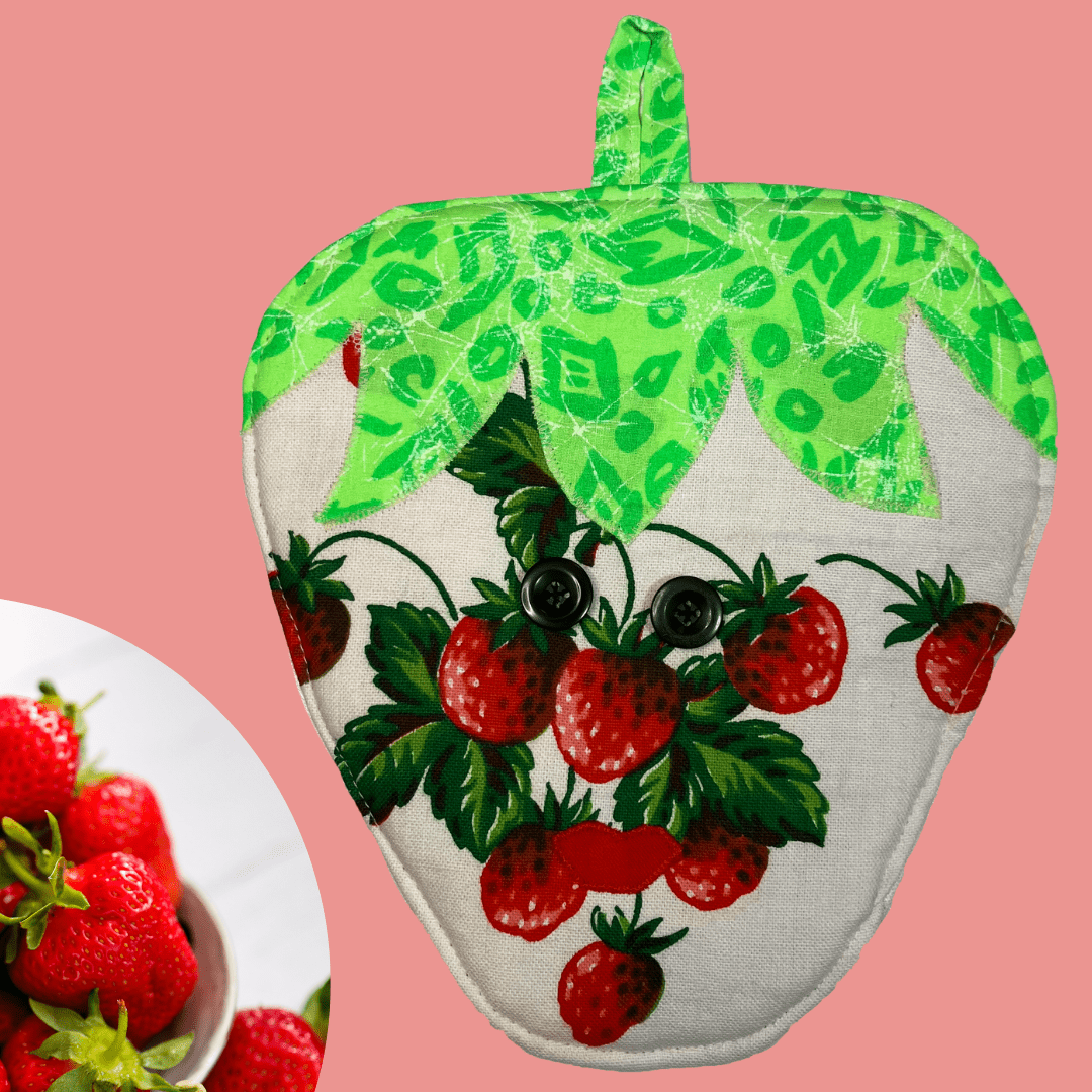 Front view of a handmade strawberry-shaped potholder with a cheerful face, featuring bright green leaves at the top and a red strawberry print with black button eyes.