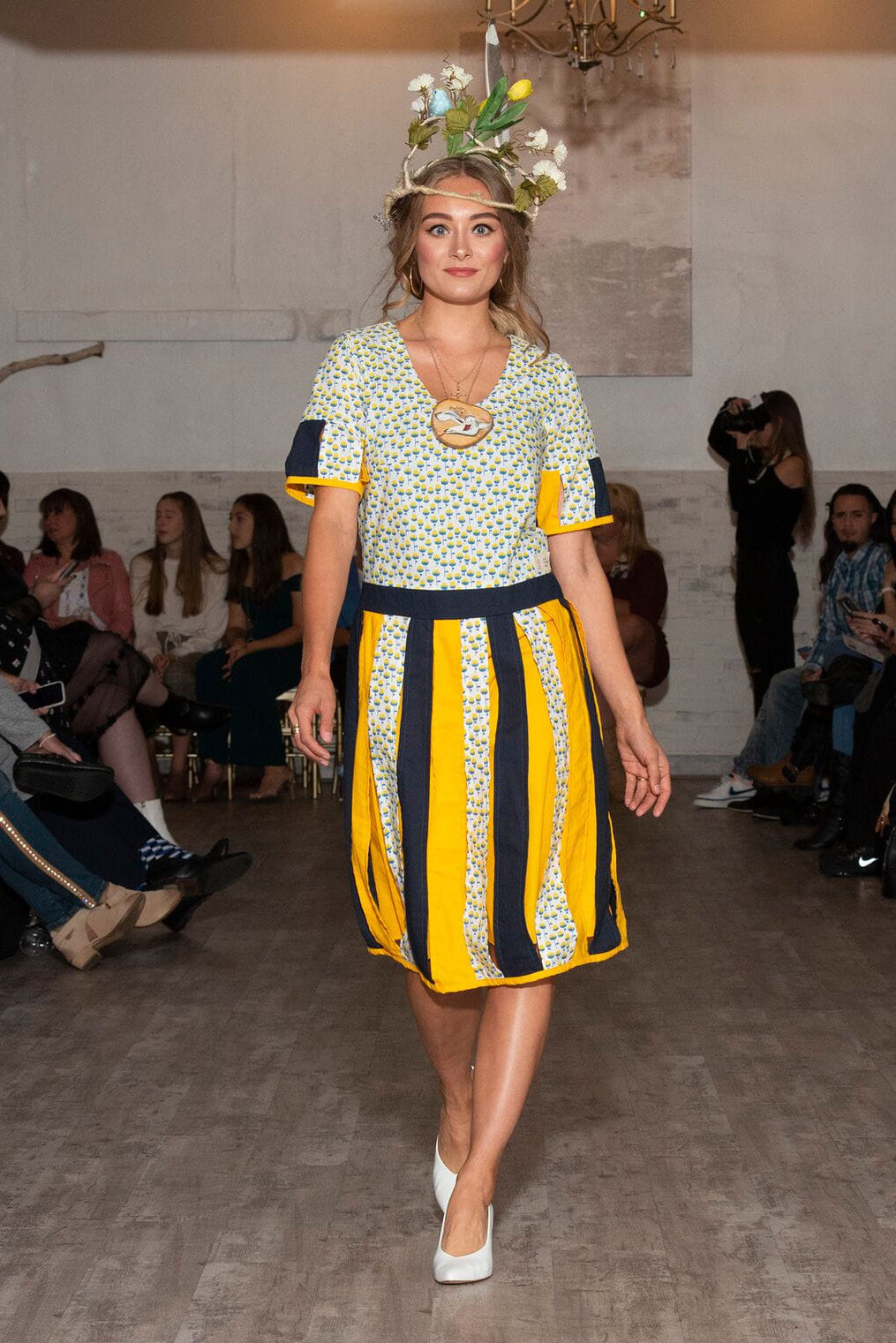 Model walking on a fashion show runway wearing a floral top and yellow and black striped skirt.