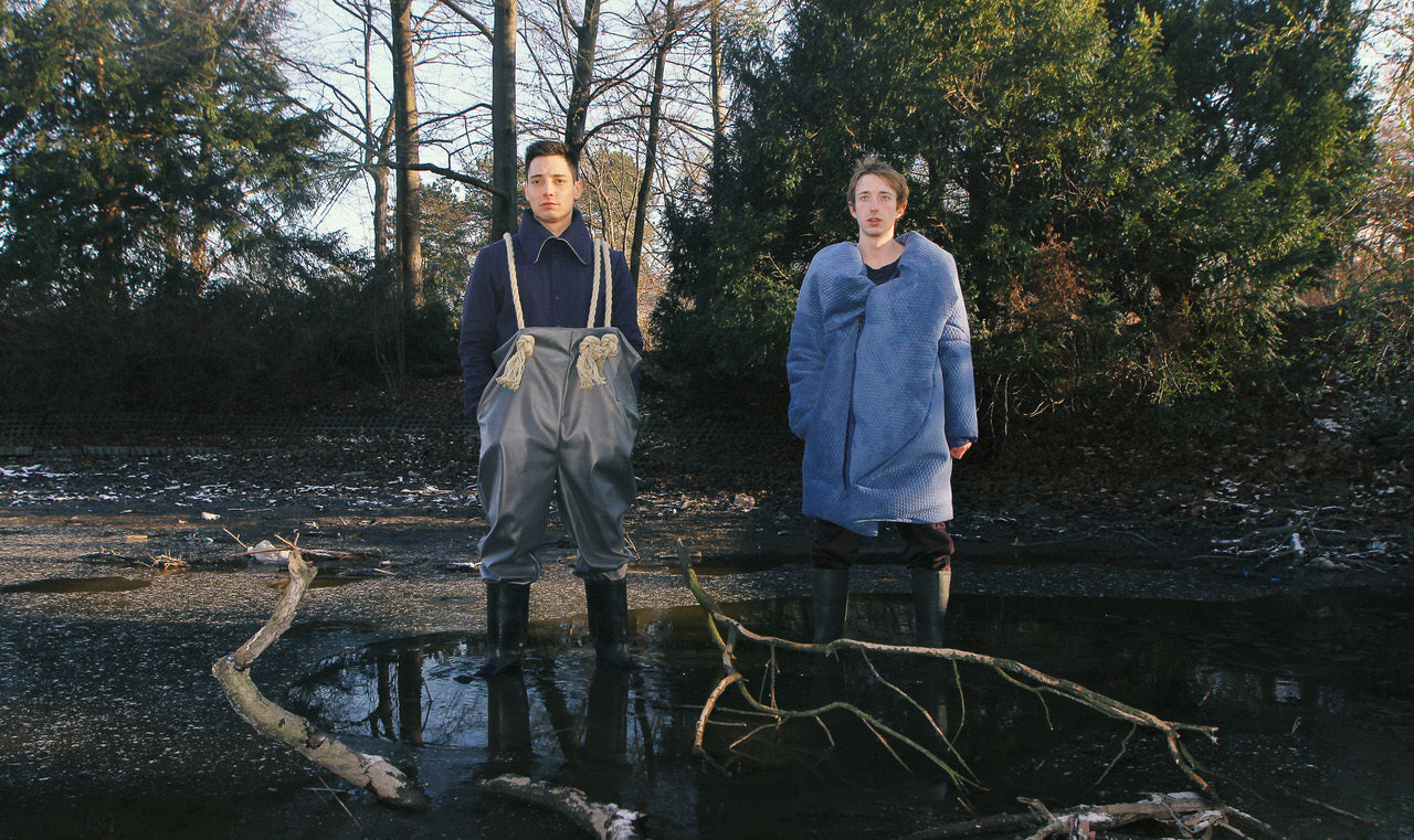 Two people standing in a forest with one wearing a blue coat and apron, the other in a light blue coat.