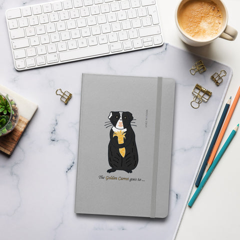 The Golden Carrot Guinea Pig Notebook resting on a desk, surrounded by a coffee mug, pencil, and keyboard, perfect for journaling and personal growth reflections designed by Anke Wonder.