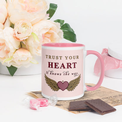 Mug with 'Trust Your Heart' quote and wings design, surrounded by flowers and chocolate.