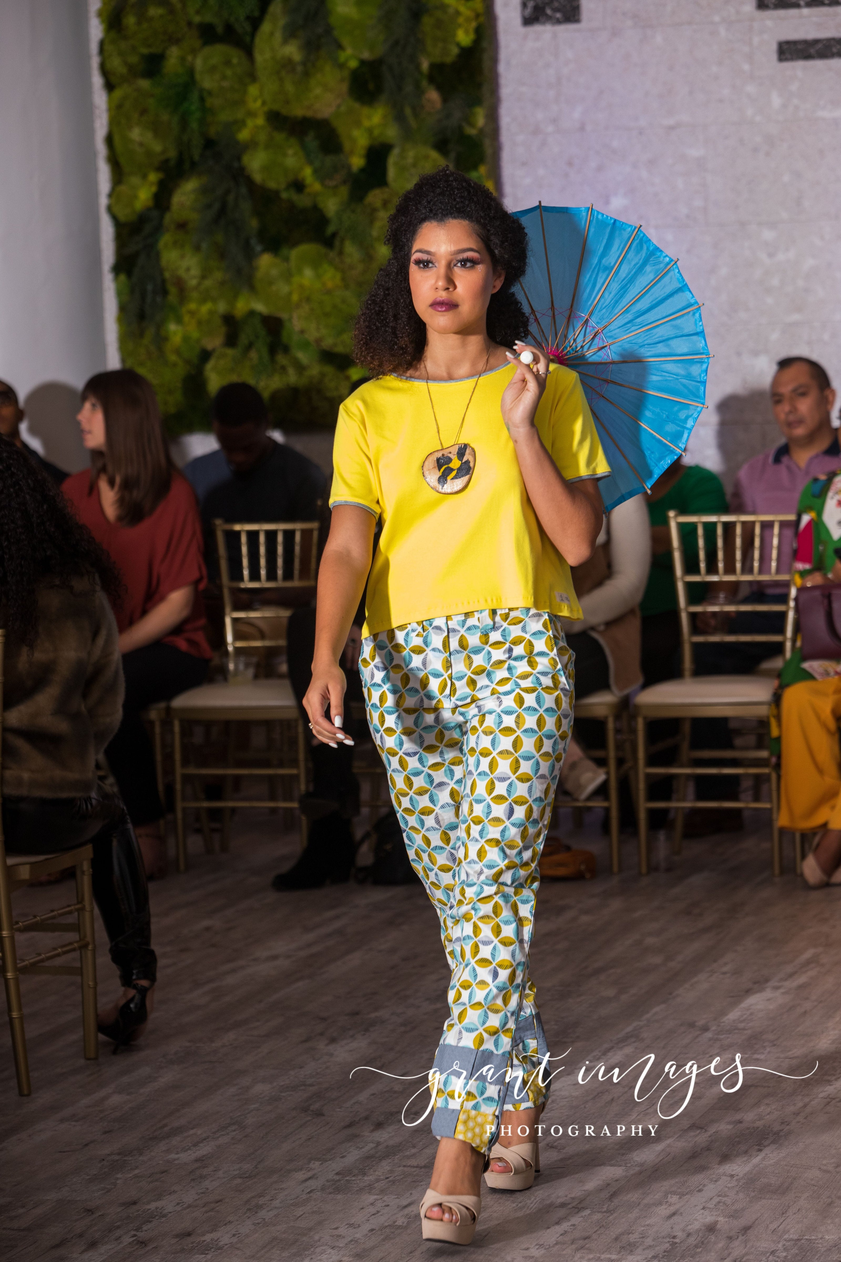 Woman in yellow top and patterned pants holding a blue umbrella at an event.