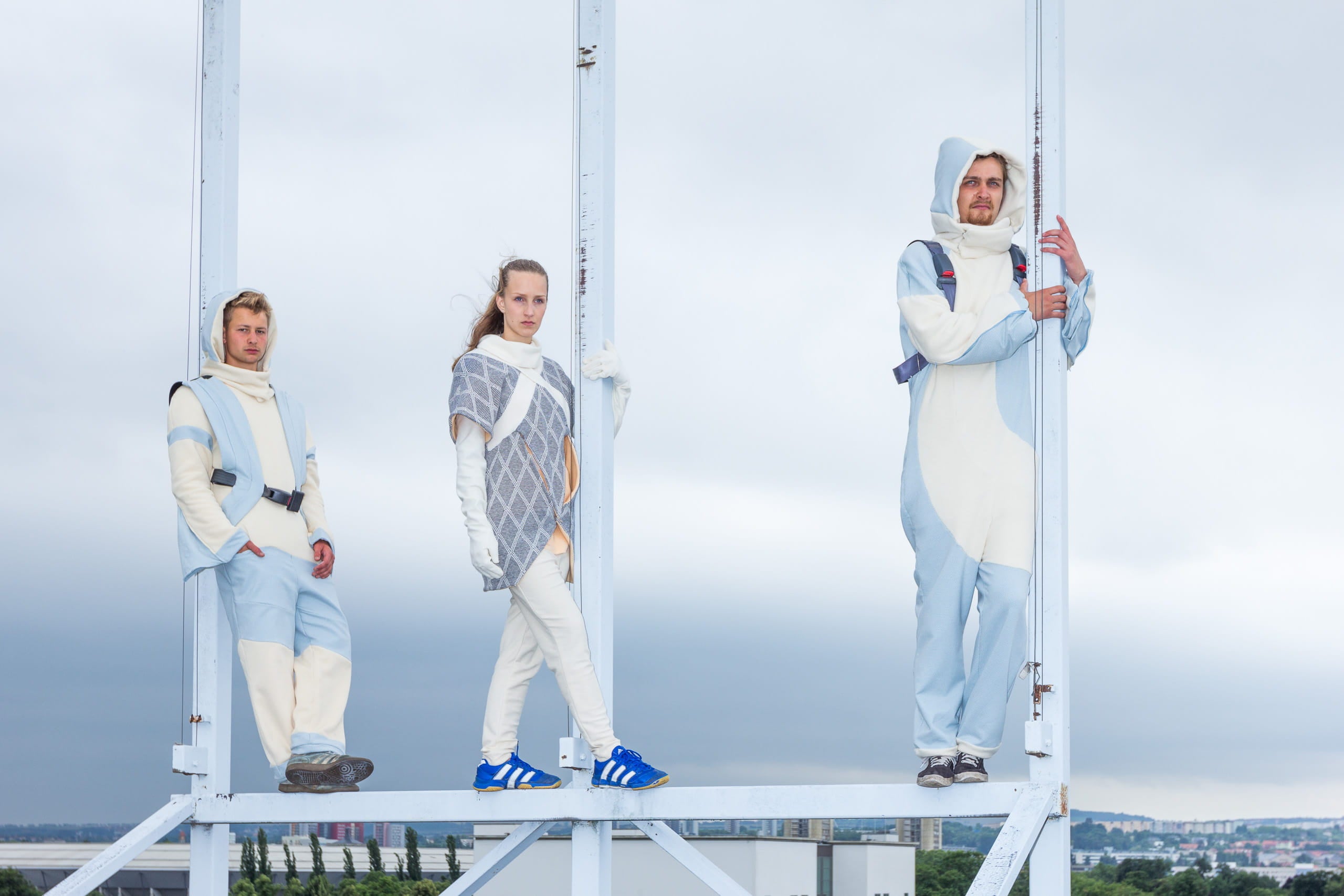 Three people wearing futuristic white suits on a metal structure with a cityscape background