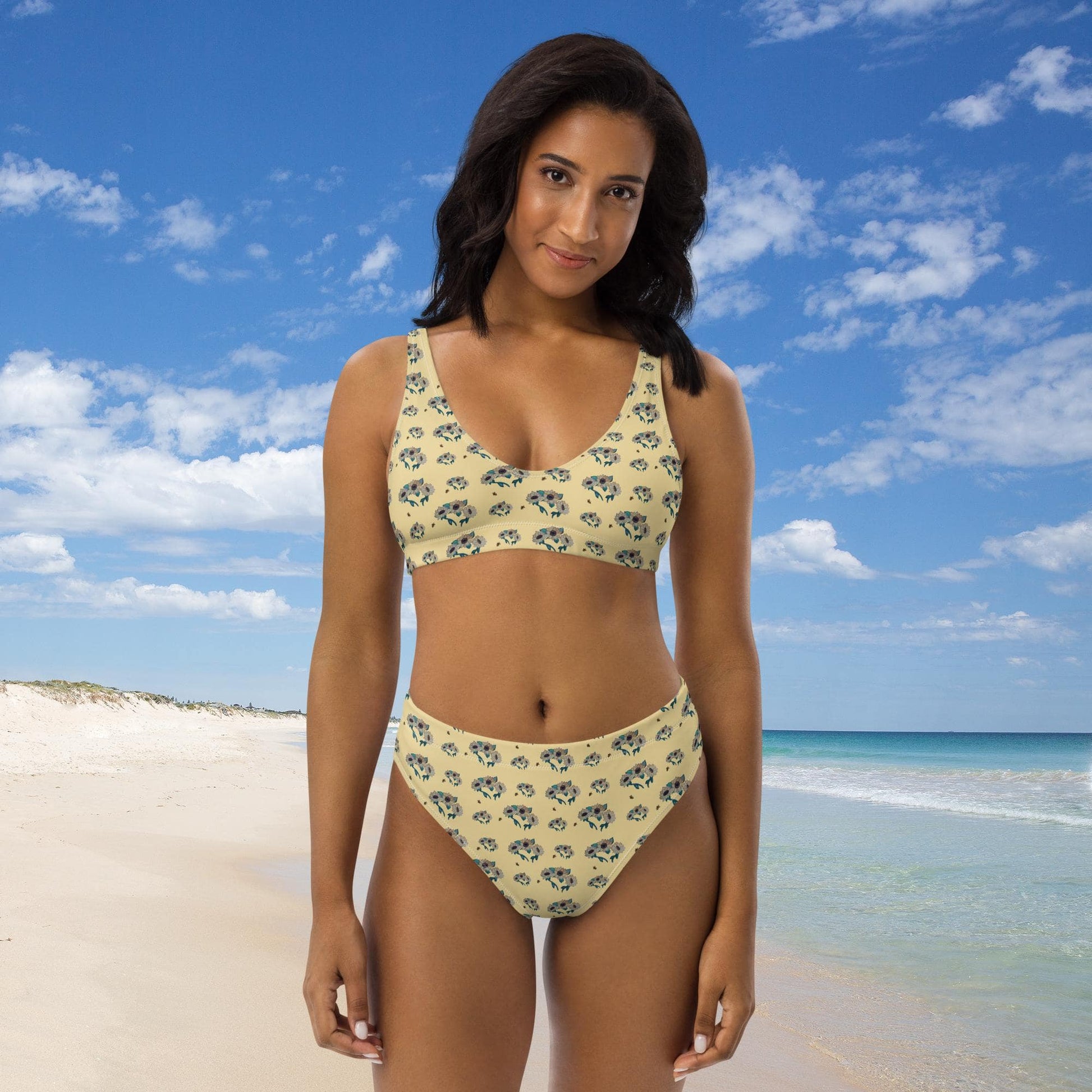 Woman in a floral sunflower bikini standing on a beach with blue sky and ocean in the background.