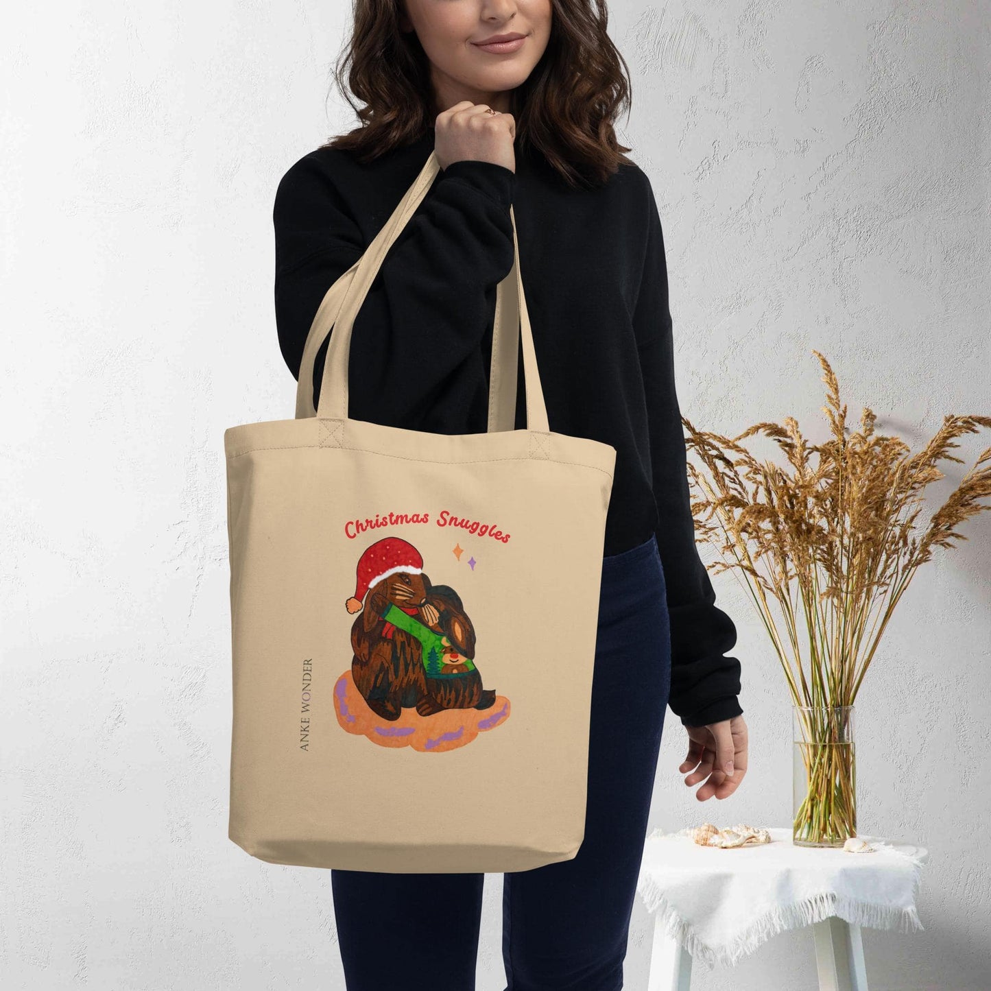 Person holding a beige tote bag with a Christmas rabbit design, standing against a white wall with a vase of dried plants.