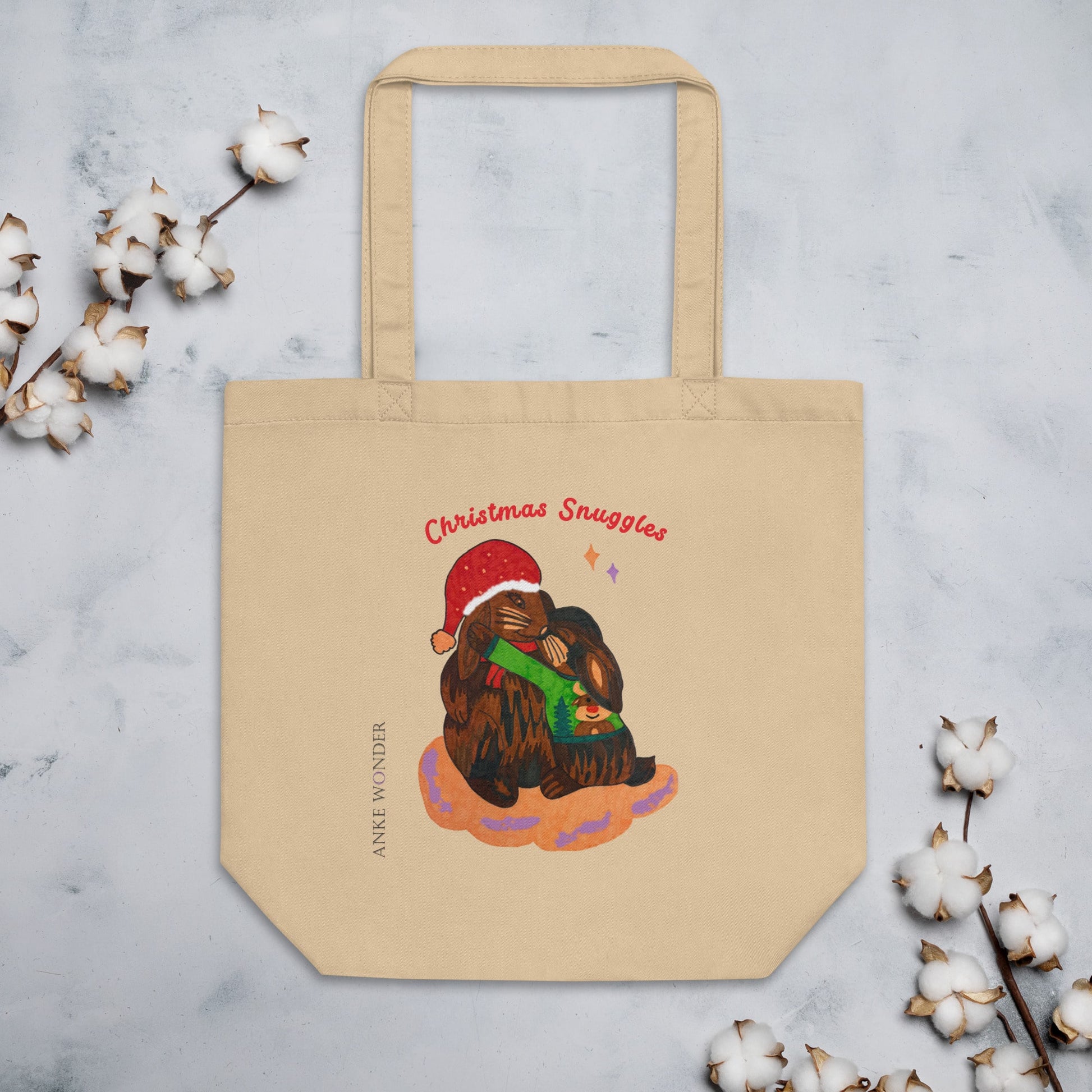 Beige tote bag with Christmas-themed design of two rabbits hugging, surrounded by cotton branches on a light gray background.
