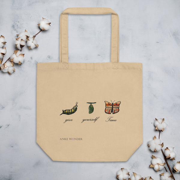 A beige organic cotton tote bag with a caterpillar-to-butterfly design and the phrase "Give Yourself Time," placed on a textured surface with cotton branches.