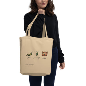 A person carrying a beige organic cotton tote bag over their shoulder, showcasing the inspirational caterpillar-to-butterfly design and the phrase "Give Yourself Time."