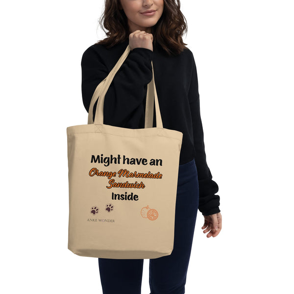 A model carries a beige organic cotton tote bag featuring a fun Paddington Bear-inspired design with text, paw prints, and an orange illustration.