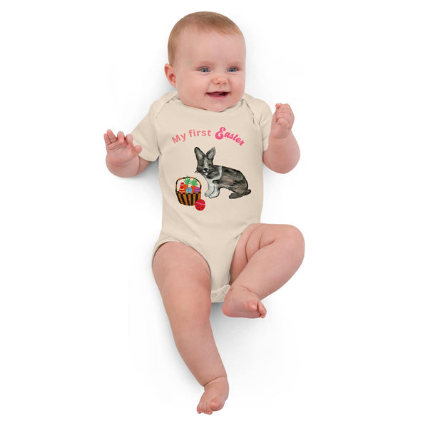 A laying baby girl wearing a short-sleeve organic cotton bodysuit with pink 'My First Easter' text, featuring a bunny and basket design by Anke Wonder.