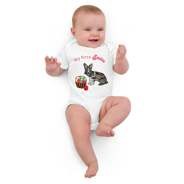 A laying baby girl wearing a short-sleeve organic cotton white bodysuit with pink 'My First Easter' text, featuring a bunny and basket design by Anke Wonder.