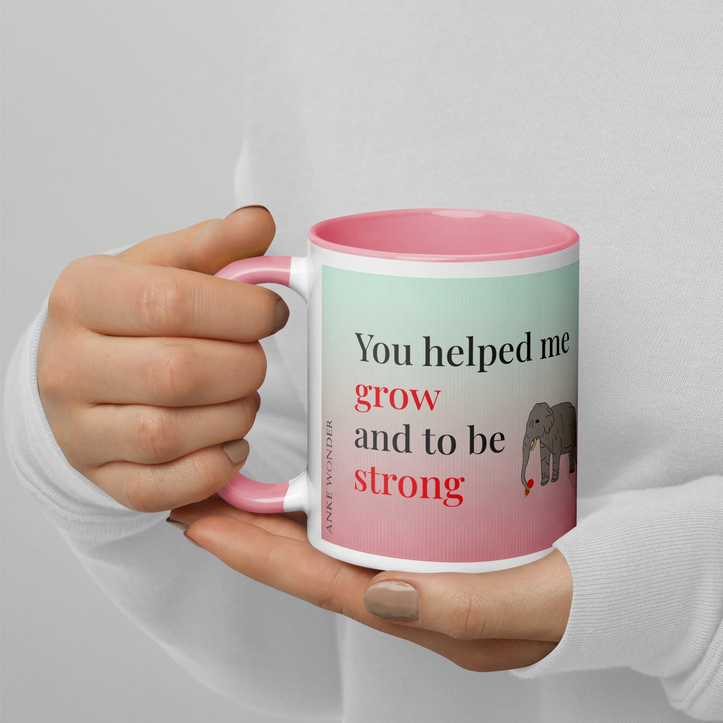 A close-up of a ceramic mug’s left side, showing the text “You helped me grow and to be strong” with a small elephant illustration. The handle and interior of the mug are pink, and the mug is being held in both hands.