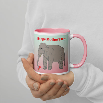 A pink-accented ceramic mug with a drawing of an elephant holding a red rose in its trunk and the phrase “Happy Mother’s Day” in red letters above. The mug is held with both hands against a light grey background.