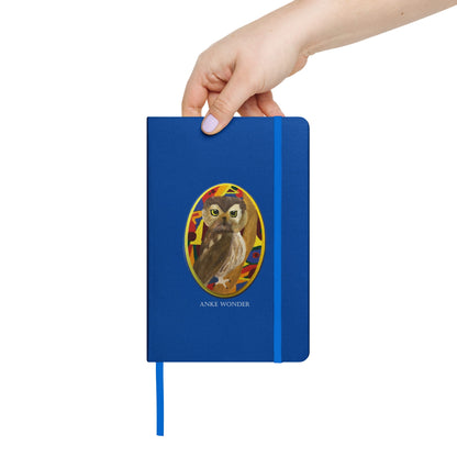 A hand holding a blue hardcover notebook with an owl portrait illustration on the front cover and a blue elastic closure, shown against a plain white background.