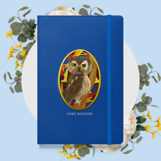 A blue hardcover notebook or journal with an oval owl illustration on the front and the text 'ANKE WONDER' below it, styled on a white circular plate with eucalyptus leaves and small yellow flowers against a light blue background.