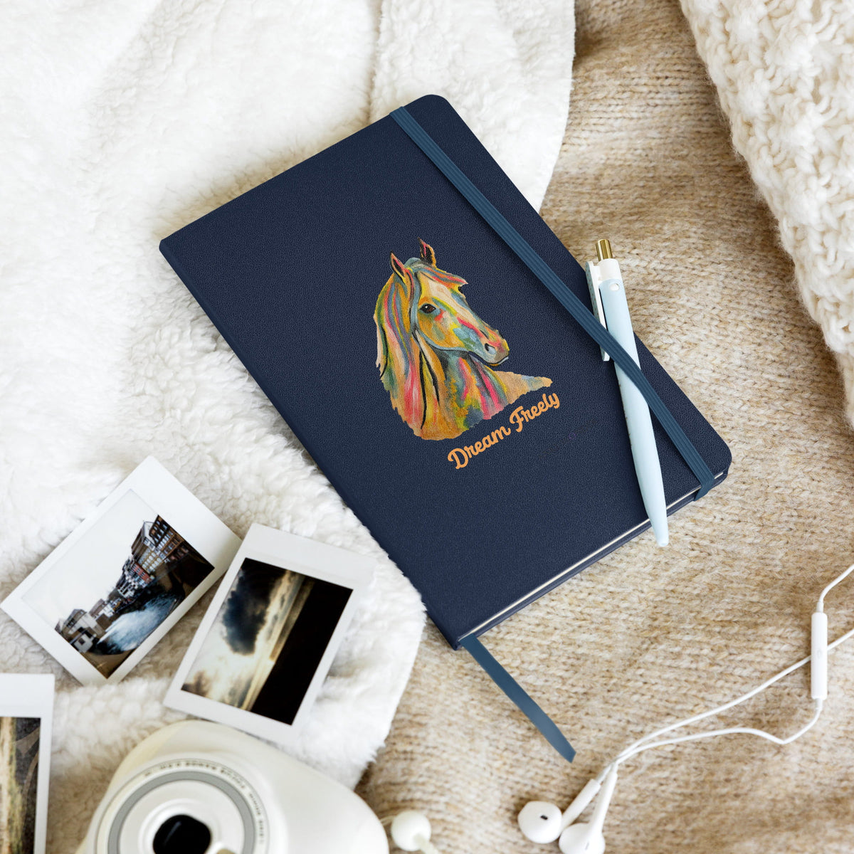 A navy blue hardcover notebook with an elastic closure, featuring a colorful painted horse illustration and the words 'Dream Ready' on the cover. The notebook is styled in a cozy flat-lay arrangement with a white pen, instant photos, earbuds, and a camera on a beige fabric background.