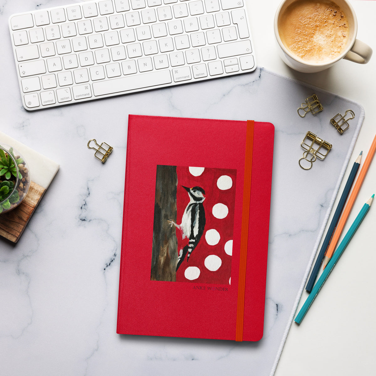 A red hardcover notebook with an elastic closure and a printed woodpecker illustration on the cover, arranged on a white marble desk with a keyboard, coffee cup, binder clips, pencils, and a small succulent.