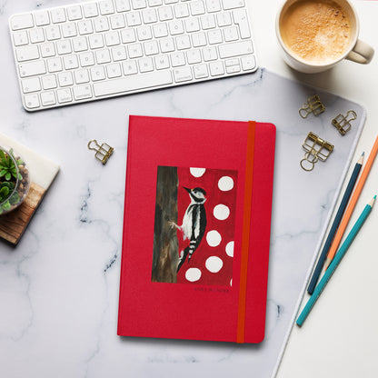 A red hardcover notebook with an elastic closure and a printed woodpecker illustration on the cover, arranged on a white marble desk with a keyboard, coffee cup, binder clips, pencils, and a small succulent.