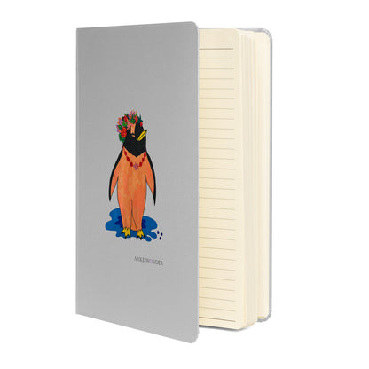 Open grey notebook with colorful artistic penguin notebook with a silver-grey hardcover, featuring a stylized orange penguin wearing a vibrant floral crown standing above a blue shadow.