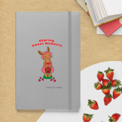 Gray notebook with a rabbit illustration and 'sharing sweet moments' text on a wooden surface with strawberries.