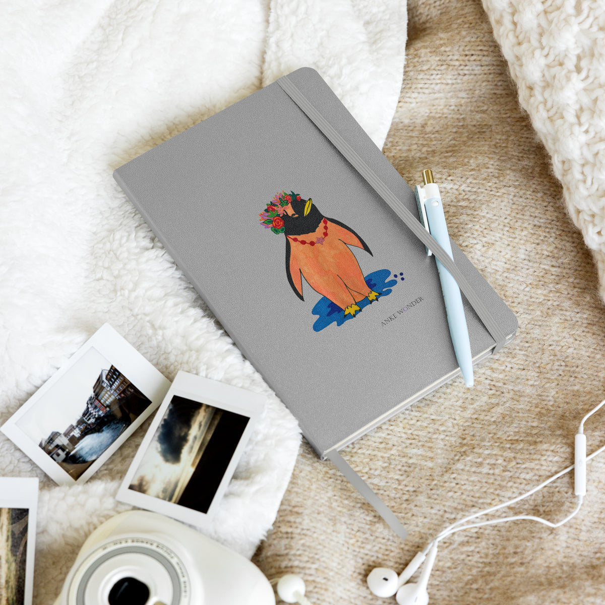 Colorful artistic penguin notebook with a silver-grey hardcover, featuring a stylized orange penguin wearing a vibrant floral crown standing above a blue shadow in a lifestlye photo.