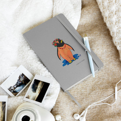 Colorful artistic penguin notebook with a silver-grey hardcover, featuring a stylized orange penguin wearing a vibrant floral crown standing above a blue shadow in a lifestlye photo.