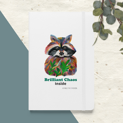 A white hardcover notebook or journal lying on a textured light surface, with a colorful painted raccoon illustration on the cover and the text 'Brilliant Chaos Inside' and 'Anne Wunder.' A teal geometric shape appears at lower left, and green leaves hang from the upper right edge.