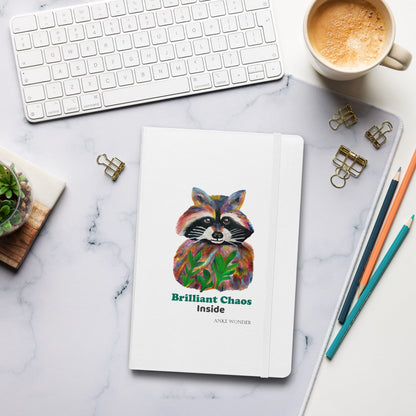 White Notebook on a desk with colorful racoon art in front and the words 'Brilliant Chaos inside'.