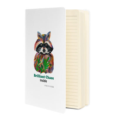 White notebook open lined with colorful racoon art in front and the words 'Brilliant Chaos inside'.