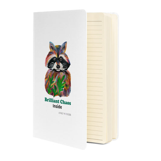White notebook open lined with colorful racoon art in front and the words 'Brilliant Chaos inside'.